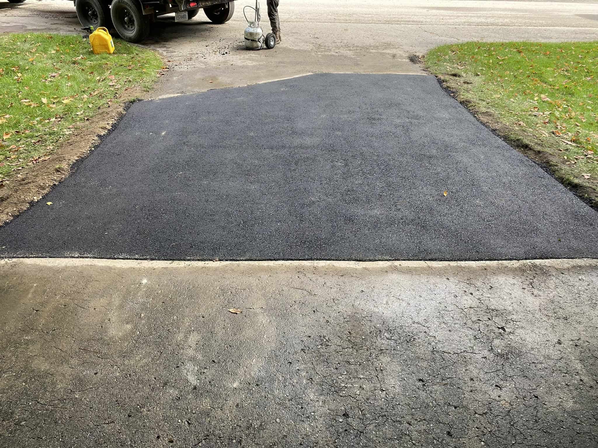 A completed structural asphalt driveway repair on Jacks Trail off Cedar Run Rd in Grand Traverse County. The image shows a large, freshly installed asphalt patch spanning the entire width of the residential driveway following the removal of a failed section, with a worker visible in the background packing up tools.