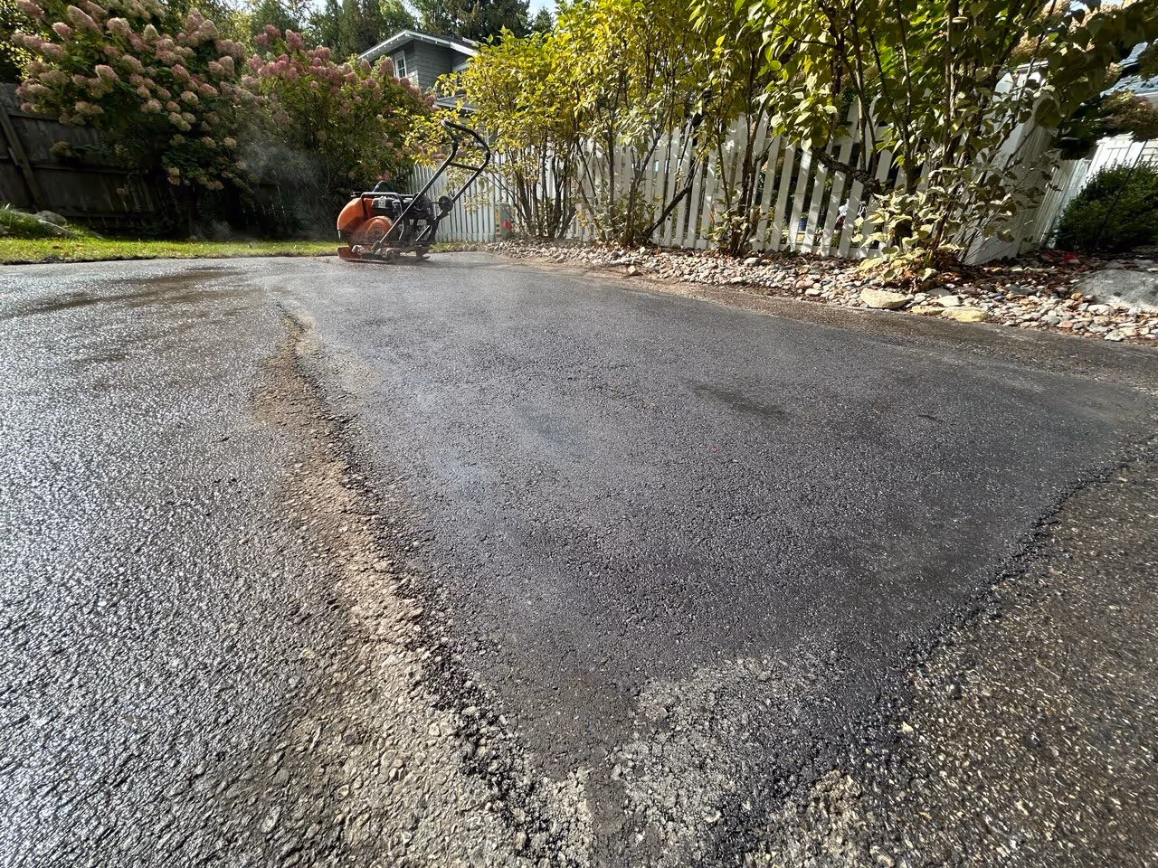 A fresh, steaming seamless infrared asphalt repair patch on a private driveway at Big Glen Lake on Dunns Farm Road in Maple City, MI, featuring a plate compactor in the background during the finishing process