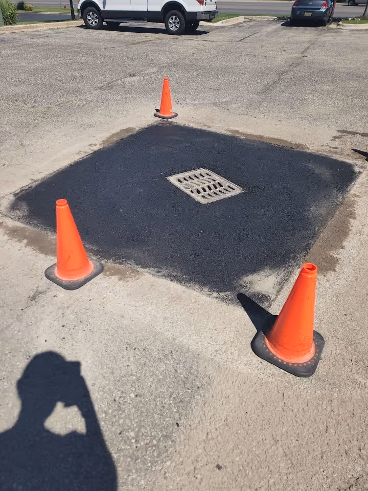 A professionally repaired parking lot catch basin with fresh, smooth asphalt at Pet Supplies Plus on South Airport Road, located in front of the Grand Traverse Mall in Traverse City, MI, secured with orange safety cones.