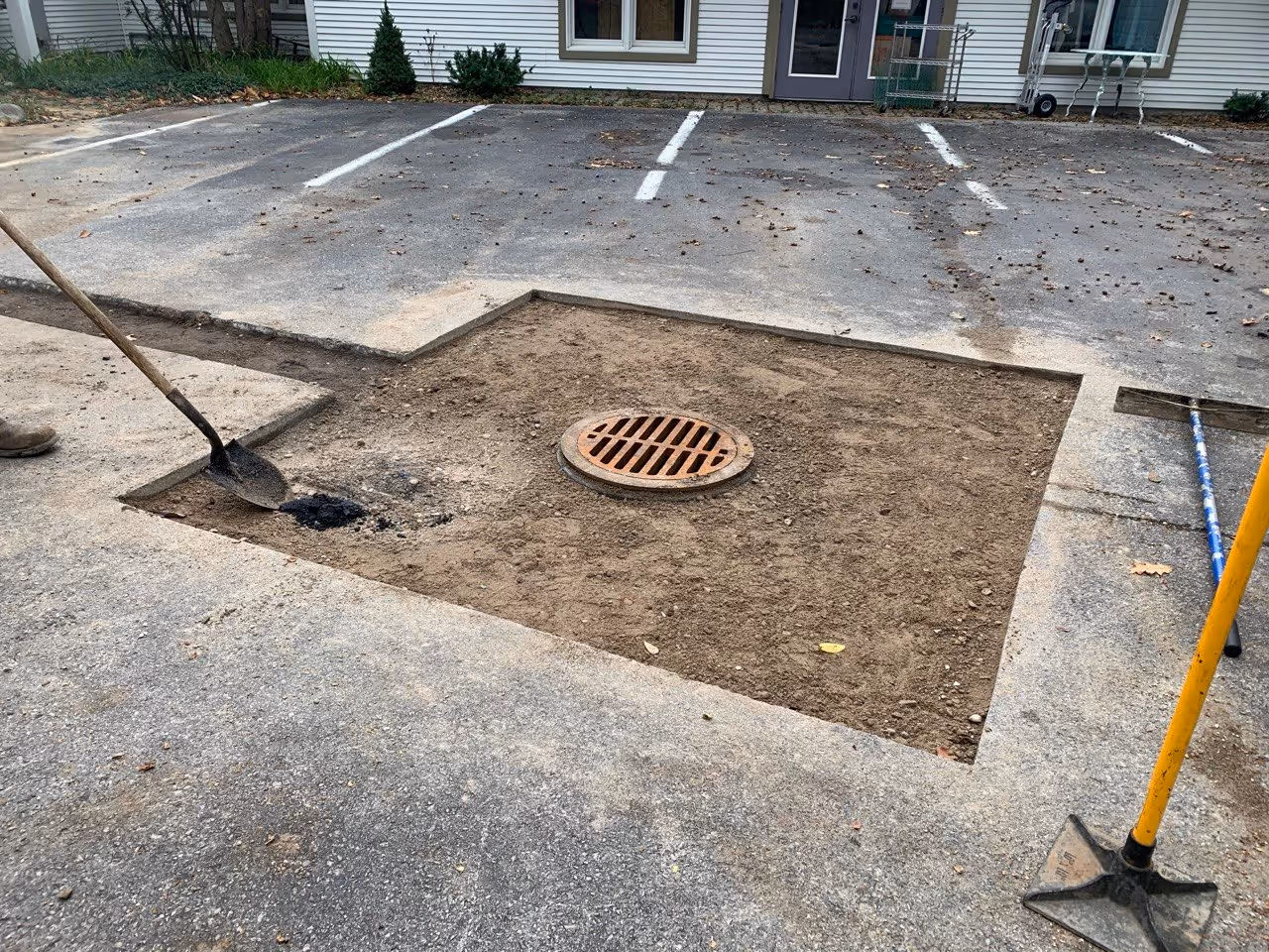 Catch basin drain installation and compacted gravel base preparation at Cottonseed Apparel on Western Avenue in Glen Arbor, MI. The area is leveled with a hand tamp and rake, prepped for professional asphalt paving.