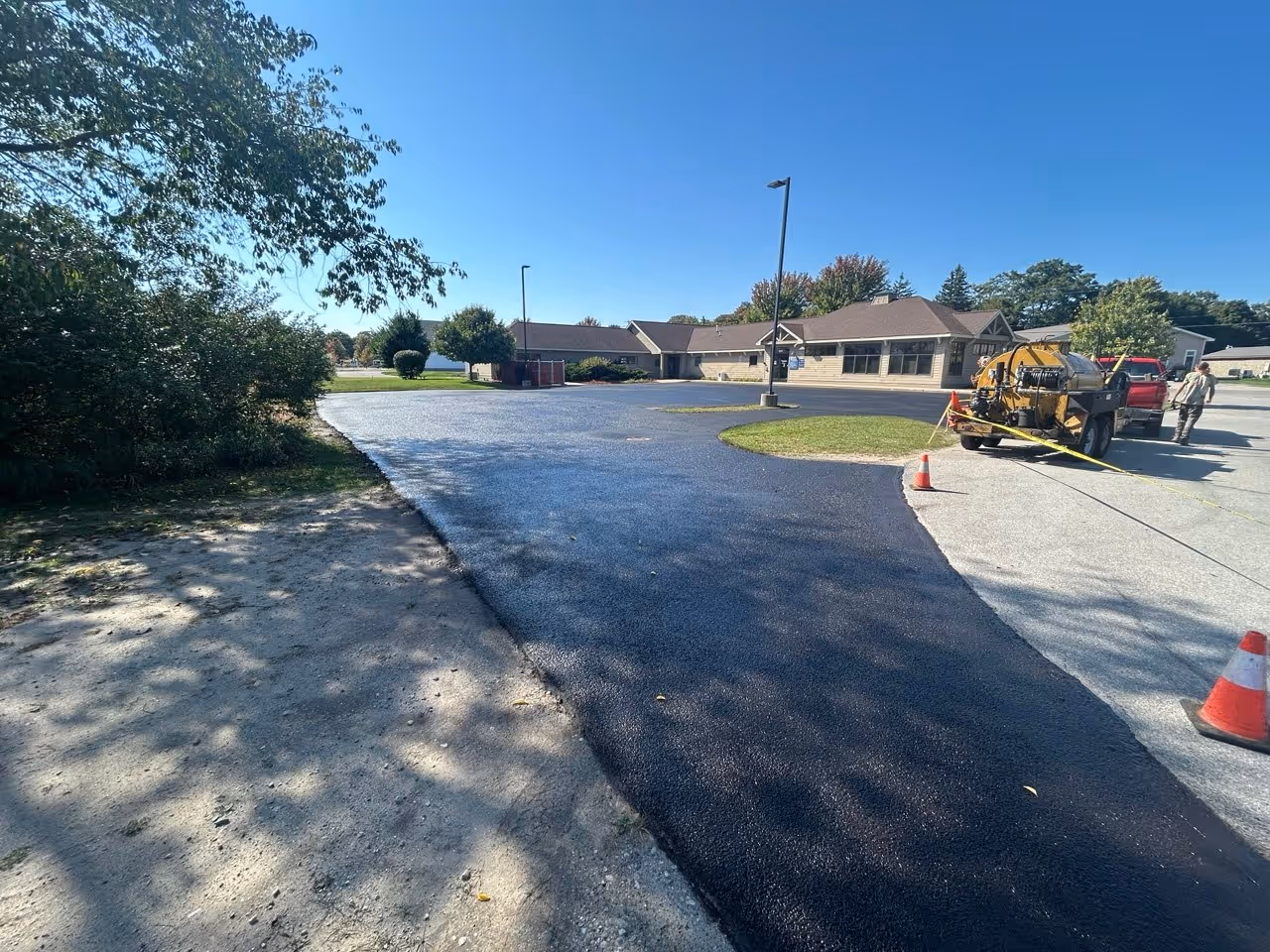 Freshly sealed asphalt parking lot at Mary Free Bed Munson Healthcare and Empire Family Care in Empire, MI on M-22. A Chevy truck and Seal-Rite 700 are visible in the background with orange cones barricading the new sealcoat.