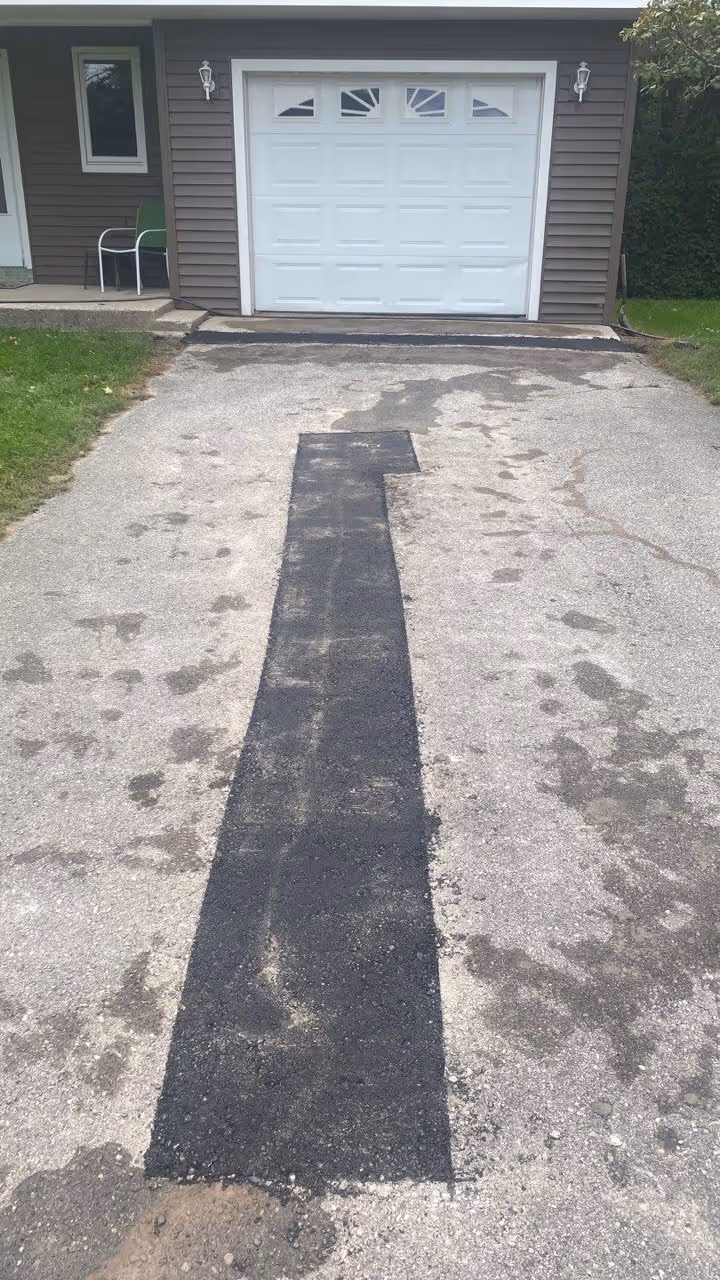 A new strip of freshly compacted asphalt patching the center of a residential driveway in Lake Leelanau, MI, providing a smooth, level surface and professional repair in Leelanau County.