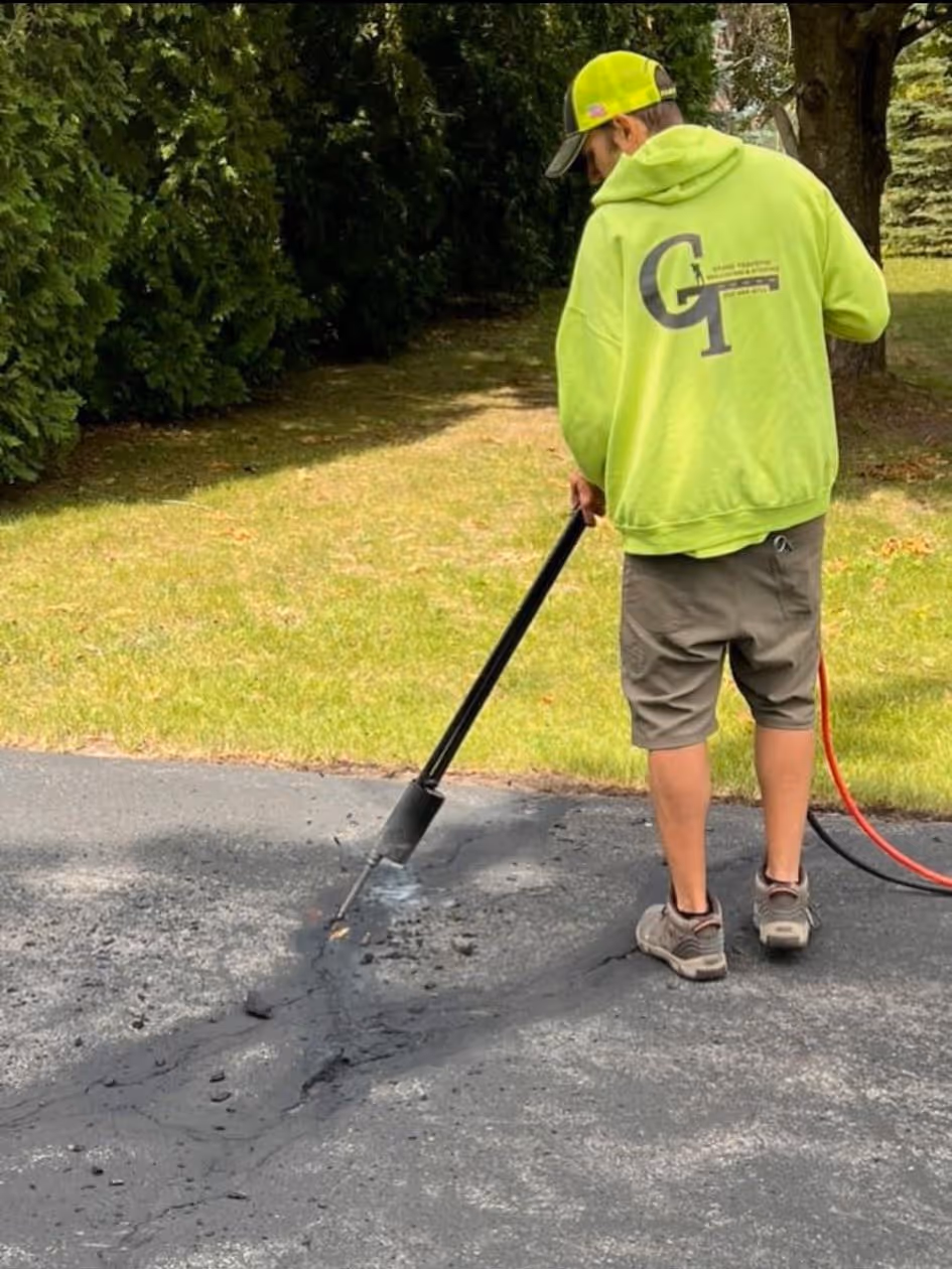 A worker in a safety green sweatshirt with the Grand Traverse Sealcoating logo uses a hot air heat lance to clean debris from asphalt cracks on a residential driveway off Cedar Run Rd in Traverse City, MI, preparing the pavement for hot rubberized crack sealing.