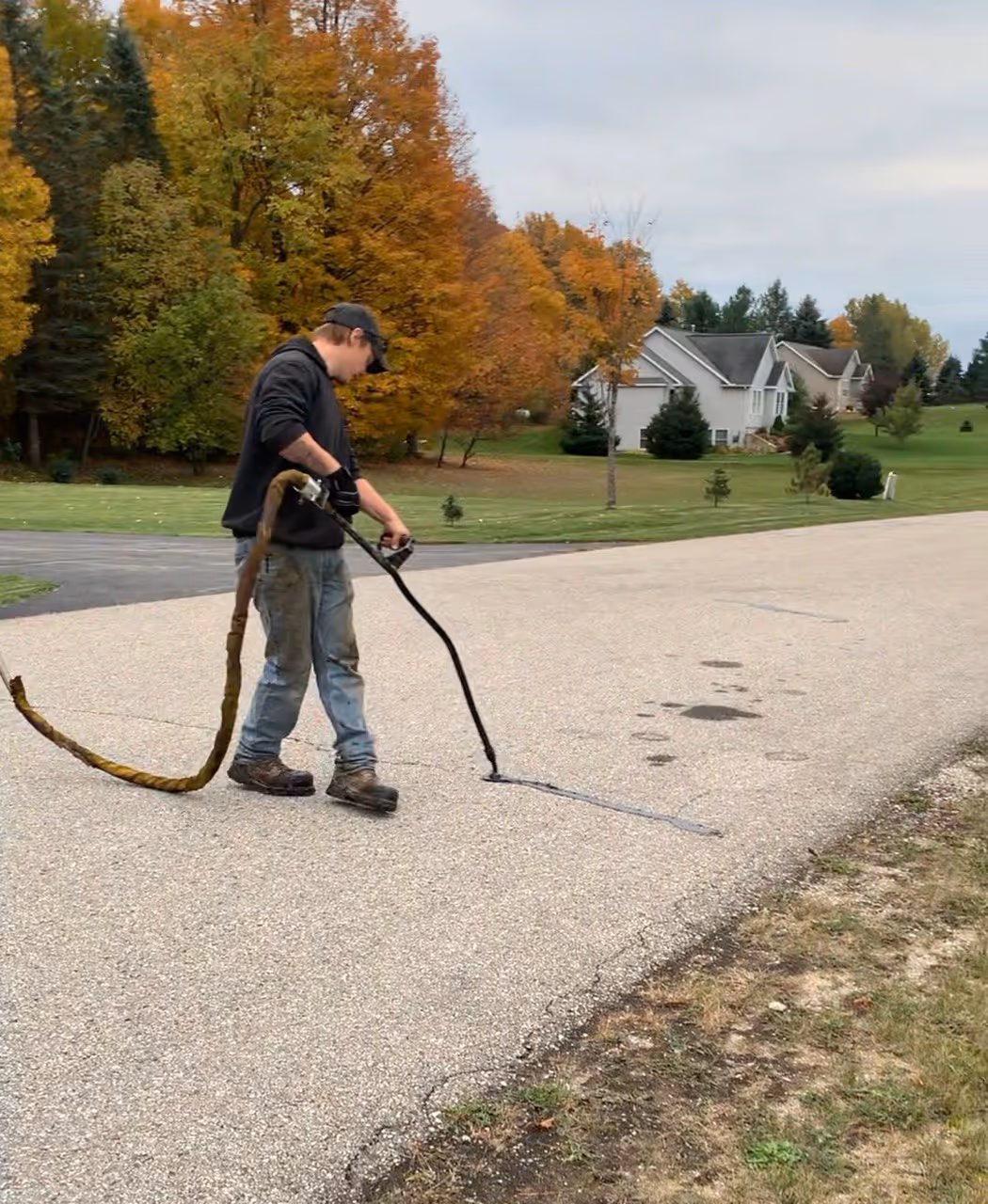 A professional applying hot rubberized sealant using a SealMaster CP-125 CrackPro wand on S Martin Lane, a private HOA road off M-72 in Leelanau County, Michigan, to prevent water infiltration and pavement damage.