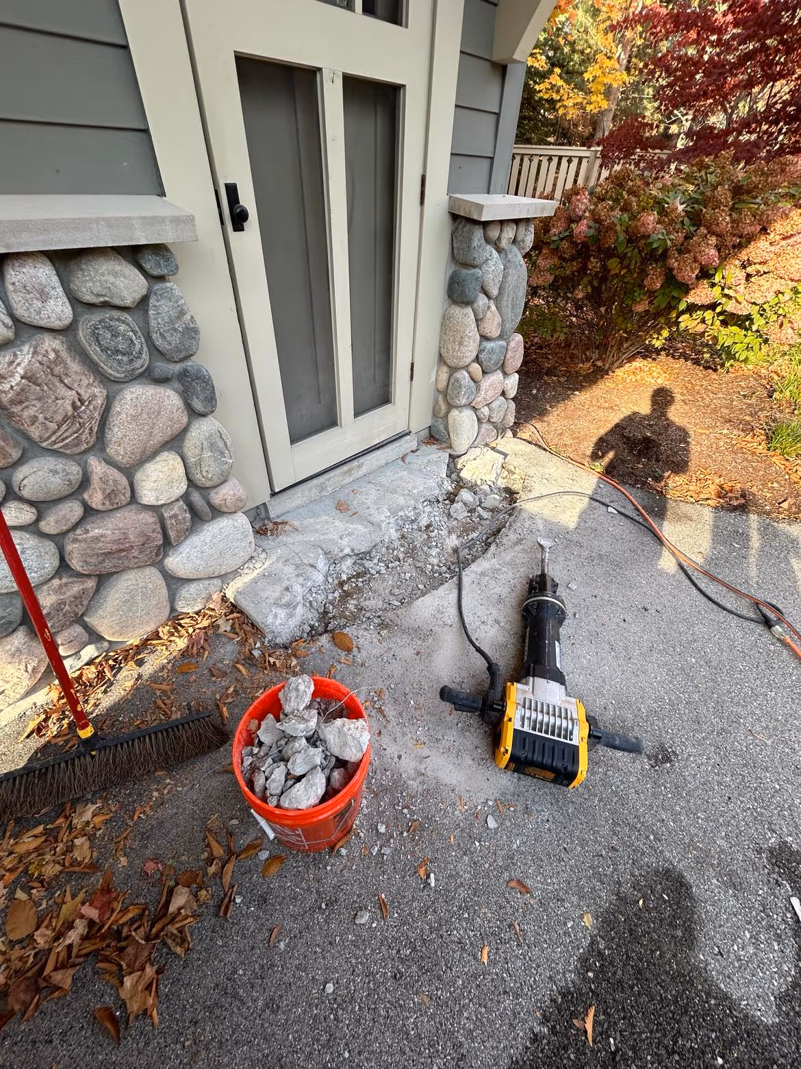 Small-scale concrete demolition in progress on S Brooks Rd in Glen Arbor, MI. A worker uses an electric DeWalt jackhammer to remove damaged concrete slabs near Big Glen Lake.