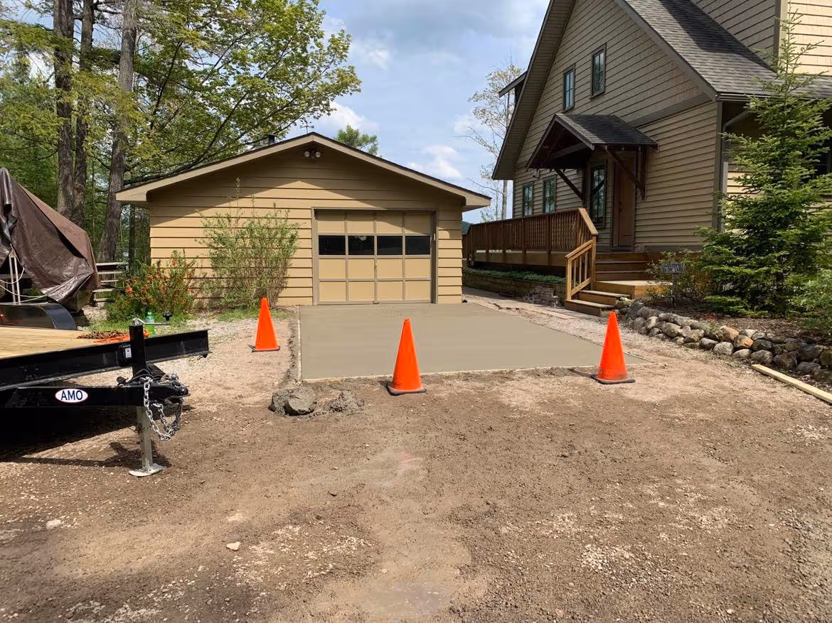 A newly poured and broom-finished concrete driveway pad for a private residence on Lime Lake, outside Maple City, MI. Three orange traffic cones block the area for proper curing.
