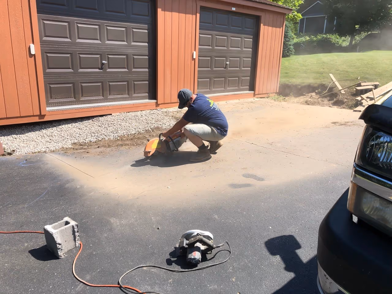 A contractor using a gas-powered asphalt saw to cut a straight, clean edge through an existing driveway to prep for a new concrete garage ramp.