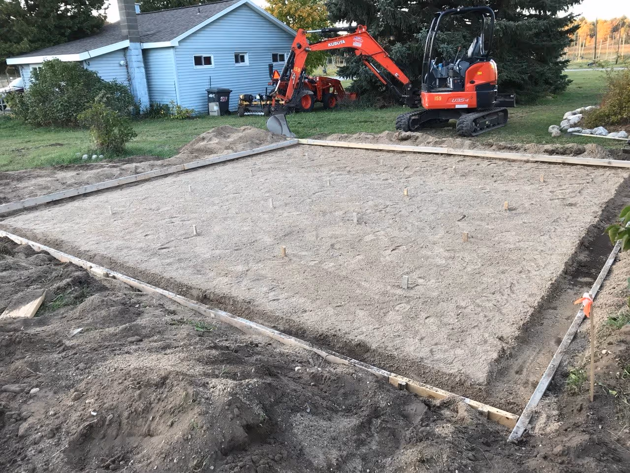 Excavating a reinforced concrete shed pad near Empire, MI, using a Kubota mini-excavator. The site is prepped with a sand base for drainage and 12-inch thickened edges for structural integrity.