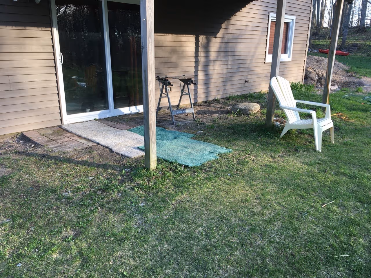 A residential backyard area in Maple City, MI, featuring rugs and lawn chairs over dirt and grass in front of a sliding glass door before a new concrete patio install.