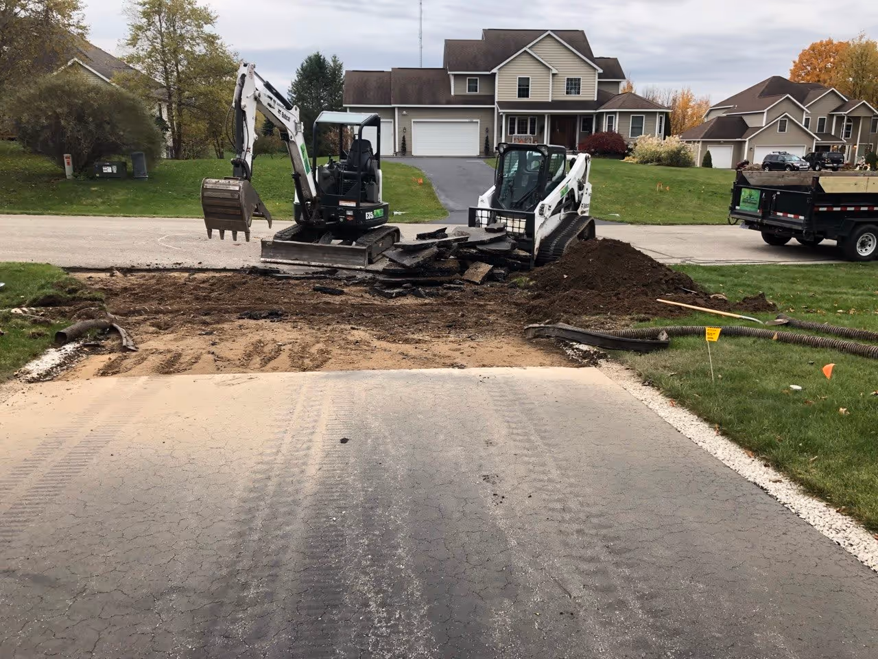 Using a skid steer and excavator to surgically remove damaged asphalt at a residential driveway in Traverse City, preparing the site for a full culvert excavation.