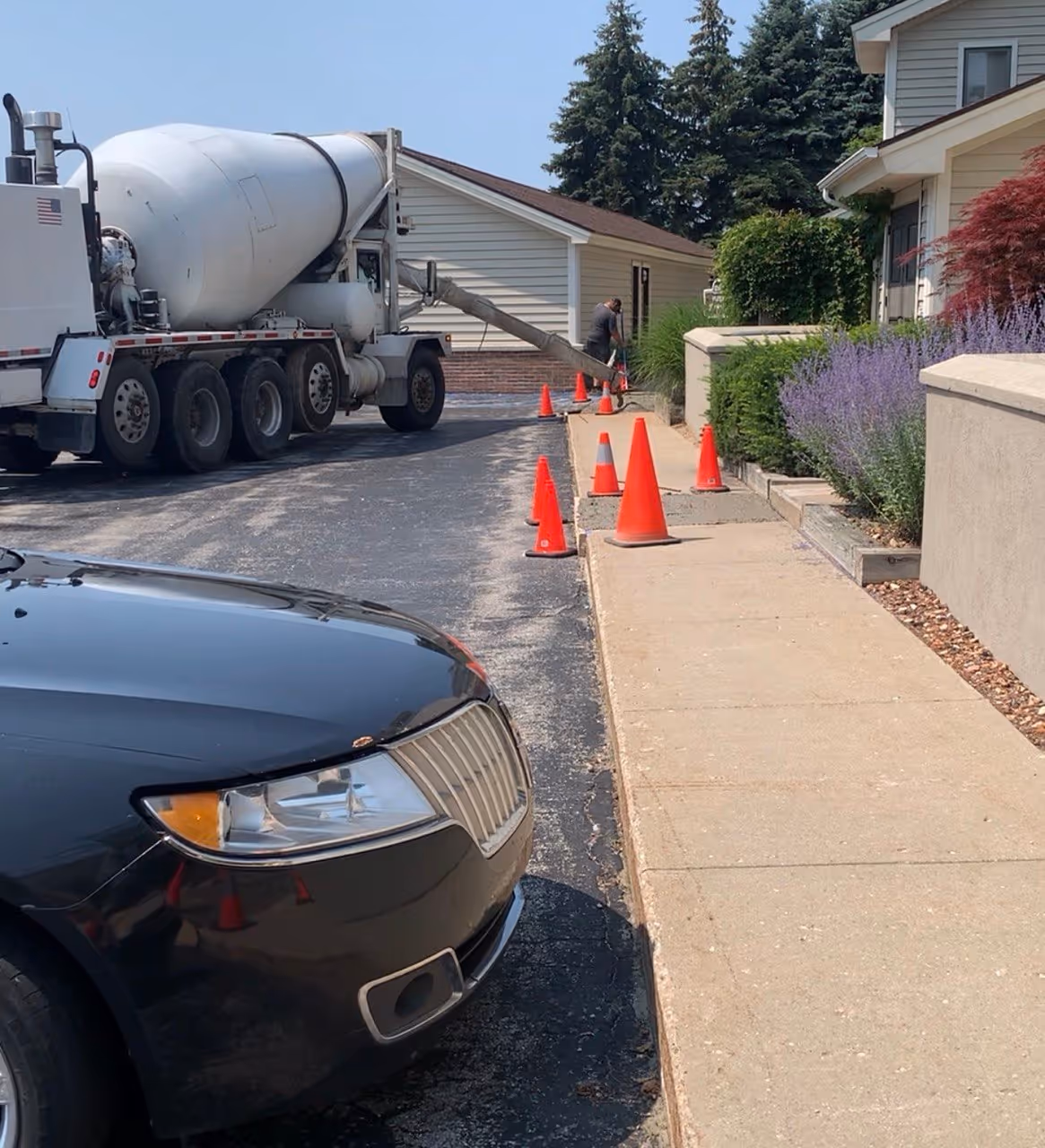 A professional concrete truck from The Concrete Service pouring and finishing new sidewalks at a waterfront condo complex in Elk Rapids, MI. Located on Dam Rd near N Bayshore Dr, the image shows freshly finished concrete sections protected by orange cones, with a worker pouring the next segment. This project, managed by Schmidt-Rogers, is situated along the Lake Michigan shoreline adjacent to Dam Beach and Fishing Park.