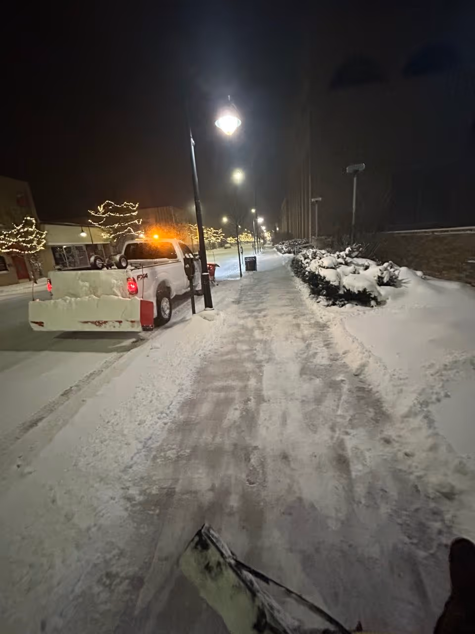 A Ford F-250 with flashing safety lights, a Boss V-plow, and a Bull Blade back blade clearing the AT&T parking lot at 142 E State Street in downtown Traverse City, Michigan. Under the glow of street lamps, the image also shows a crew member hand-shoveling the sidewalk to ensure safe pedestrian access for this high-traffic commercial location.