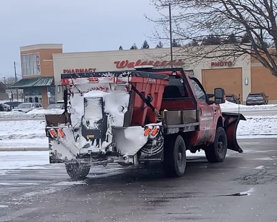 A Ford F-350 truck plowing the Chase Bank parking lot in Traverse City. The photo captures the Boss V-plow clearing lanes and the VBX 8000 spreader applying salt for ice control. In the background, the Walgreens sign at Chums Corners marks our high-visibility service area in Northern Michigan.