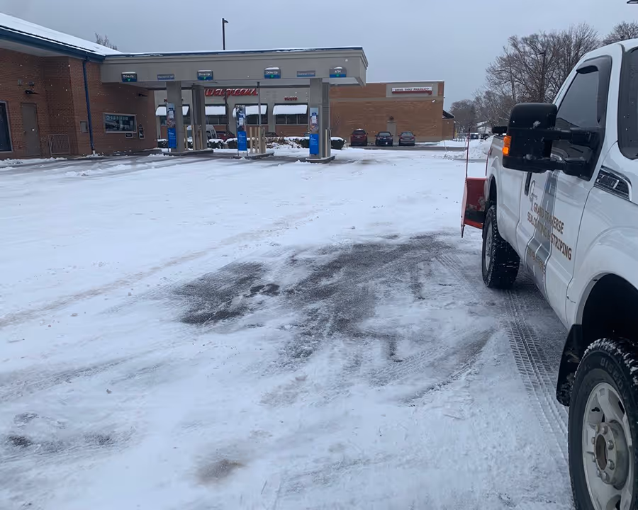 A Grand Traverse Sealcoating & Striping Ford F-250, featuring a Bull Blade back blade, performs precision snow removal at the Chase Bank drive-up teller windows in Traverse City. Located at the busy corner of W South Airport Rd and N Garfield Rd, the image showcases our specialized equipment clearing tight commercial lanes to ensure early morning banking access.