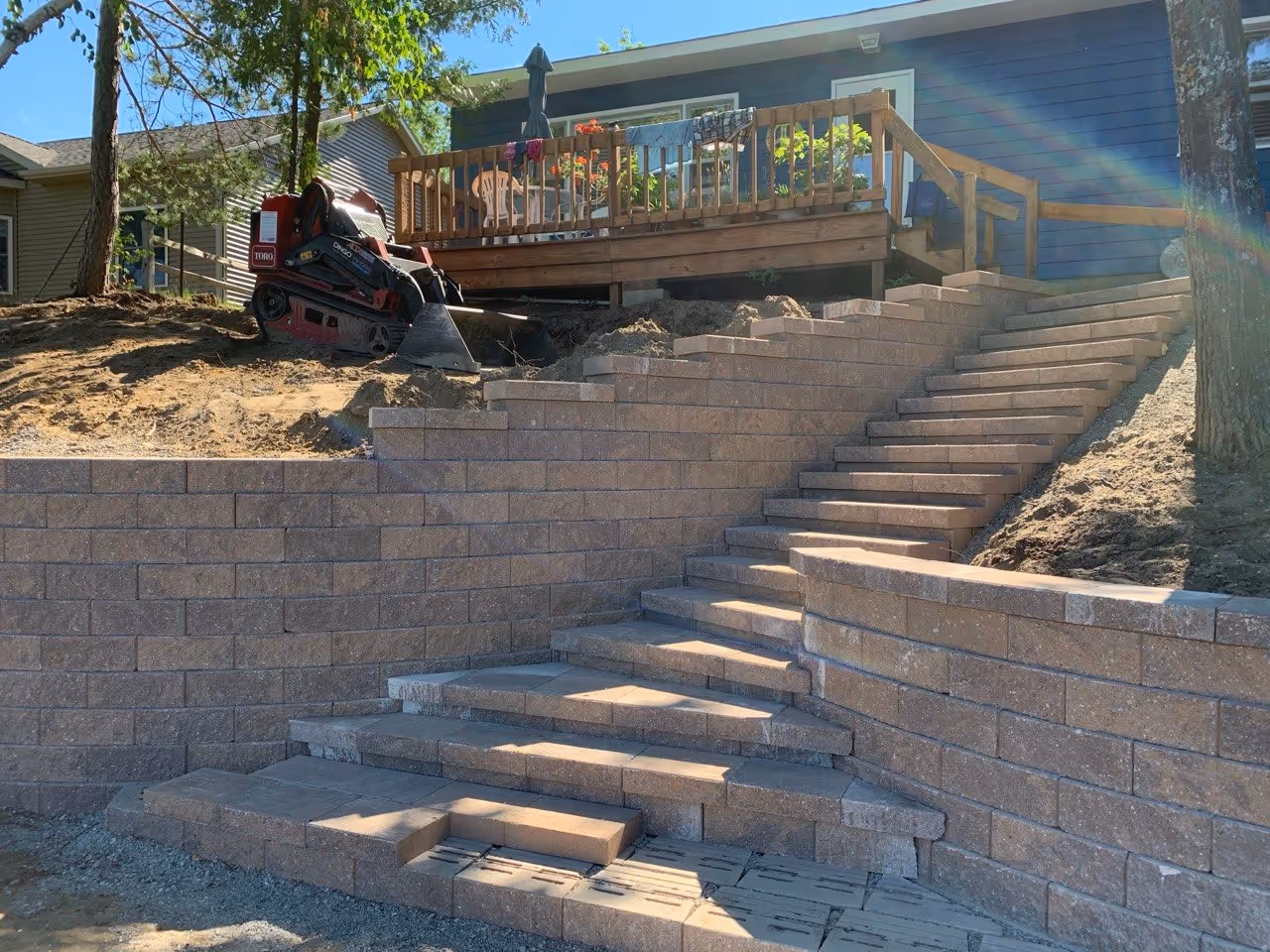 Installing smooth Versa-Lok wall caps to finish the staircase. A Toro mini skid loader is visible in the background, assisting with final material placement on the Silver Lake job site.