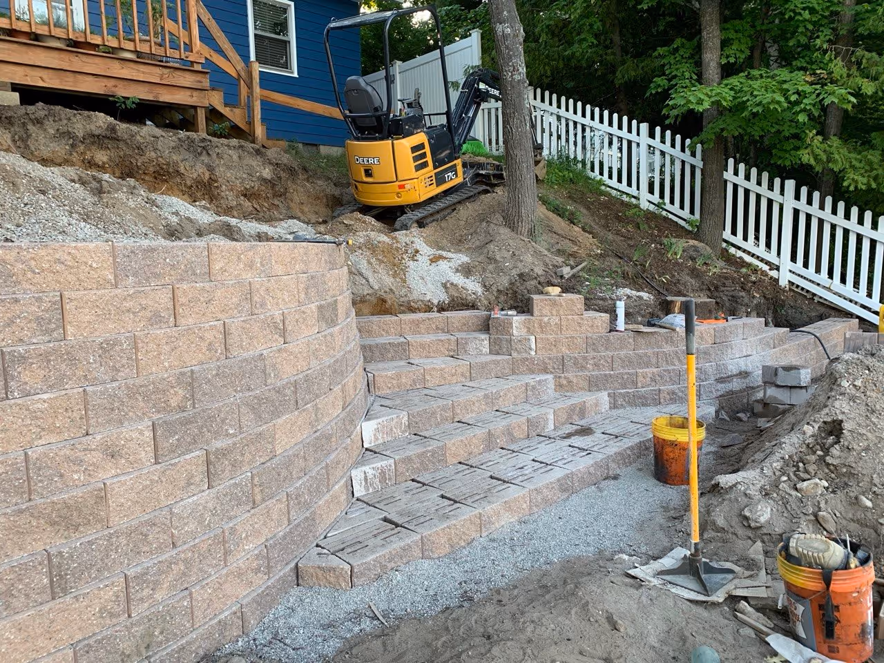 Building the Versa-Lok stairs and walls on a steep hillside. A John Deere mini excavator performs precision grading to ensure the structural integrity of the tiered wall system.