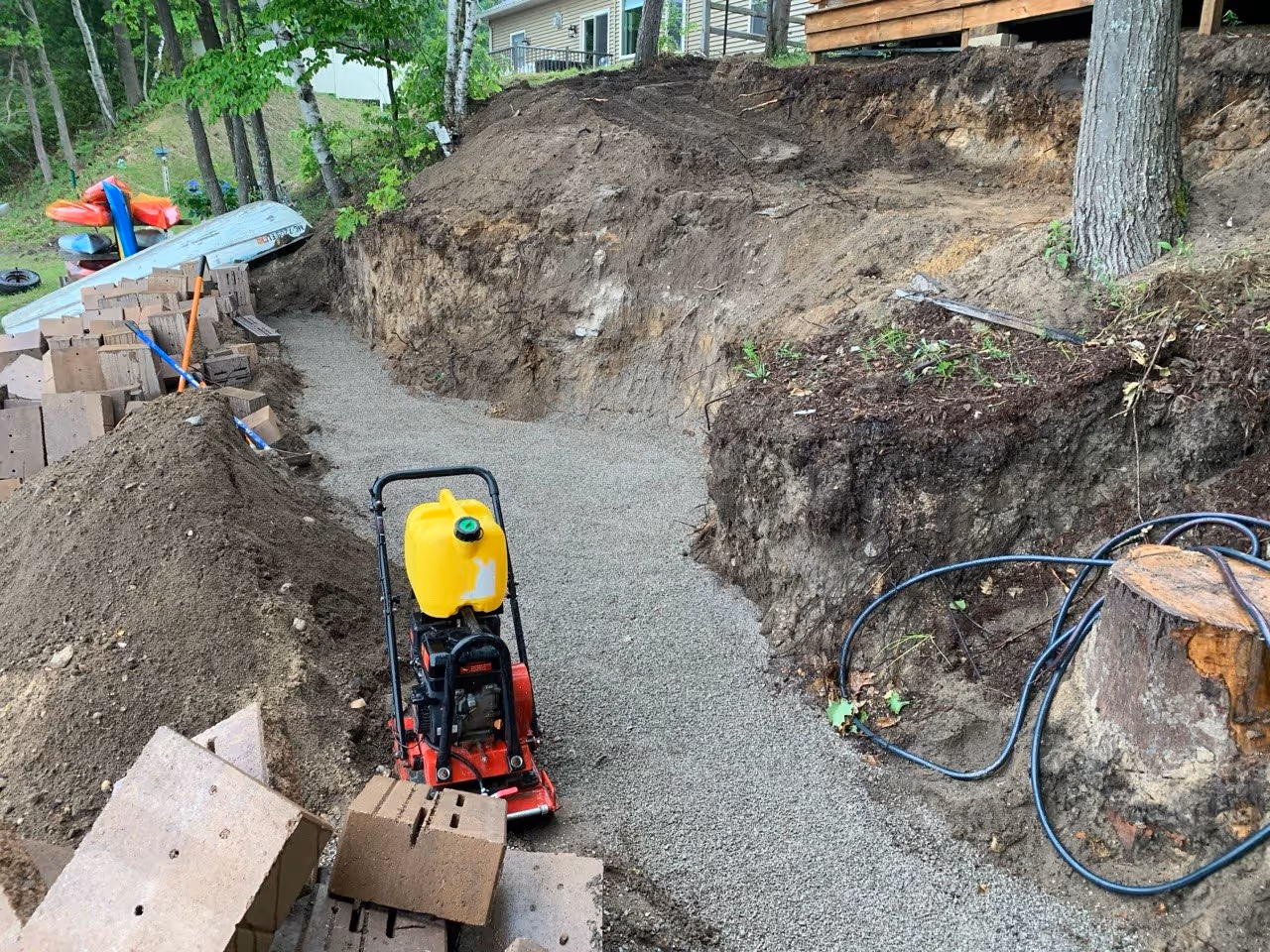 A plate compactor preparing the fresh stone base. This compacted foundation is critical for preventing wall settling and ensuring the longevity of the Versa-Lok installation in Northern Michigan.