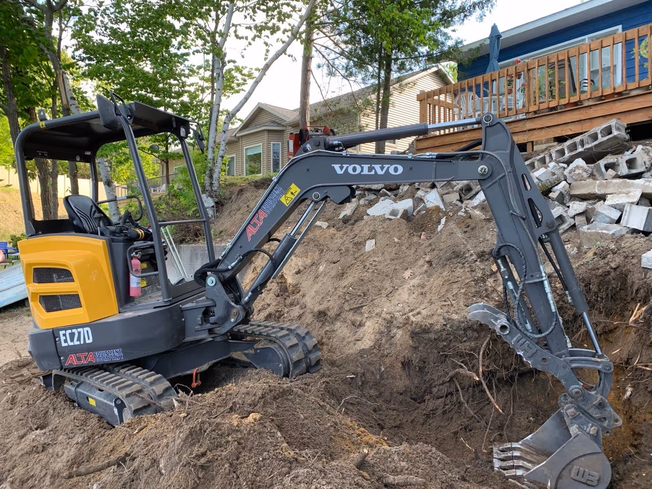 A Volvo mini excavator digging stone foundation footings on what will become the beach area. This stage anchors the entire shoreline stabilization project.