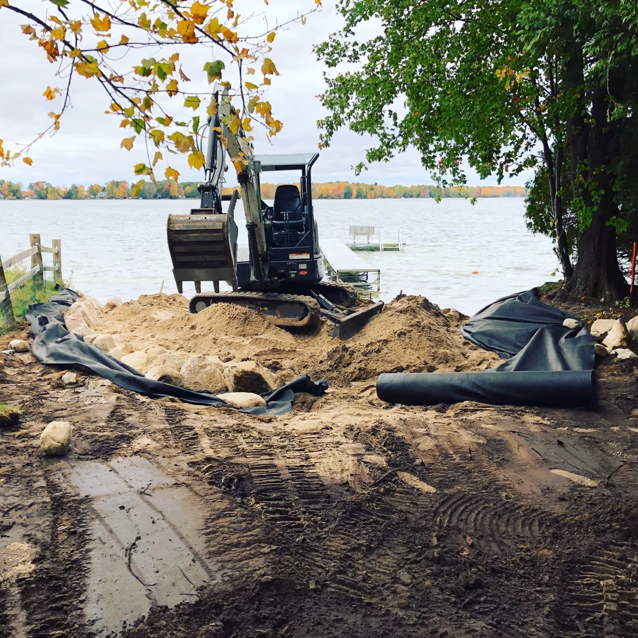 Installing a new beach on Green Lake. A Bobcat mini excavator spreads sand over heavy-duty landscape fabric, with a silt fence installed to protect the water quality during construction.