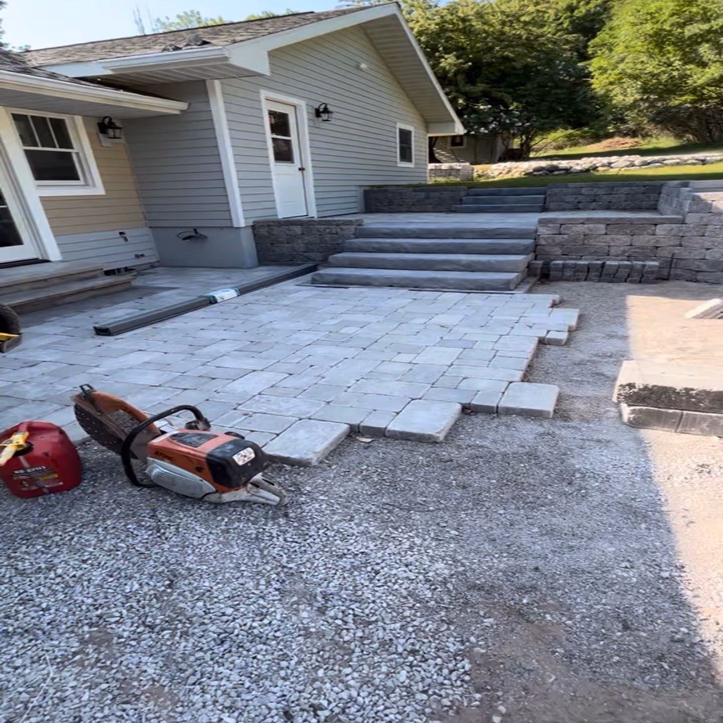 A professional hardscape construction site in Frankfort, MI, featuring a STIHL Cutquik TS 800 Cut-Off Saw and mixed gas can ready for precision paver cutting. The image shows a patterned paver patio halfway installed on a compacted crushed stone base, surrounded by newly built structural retaining walls for a waterfront property in Northern Michigan.