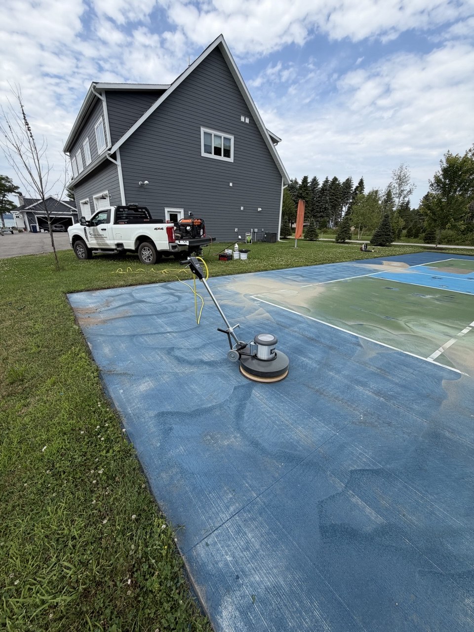 Technician using a Clarke floor sander with a sandpaper driver attachment to strip peeling paint layers from a concrete pickleball court on Torch Lake, MI.