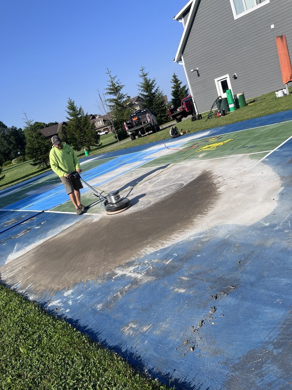 Worker running a Clarke floor sander over a repaired area patched with SportMaster acrylic patch binder, Portland cement, and sand to smooth and blend it with the concrete court.