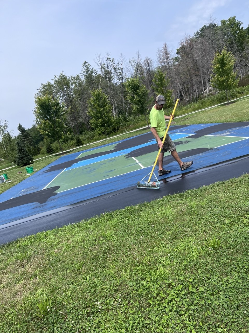 Worker using a professional squeegee to spread the first heavy sand load coat of black SportMaster acrylic resurfacer across the court surface.