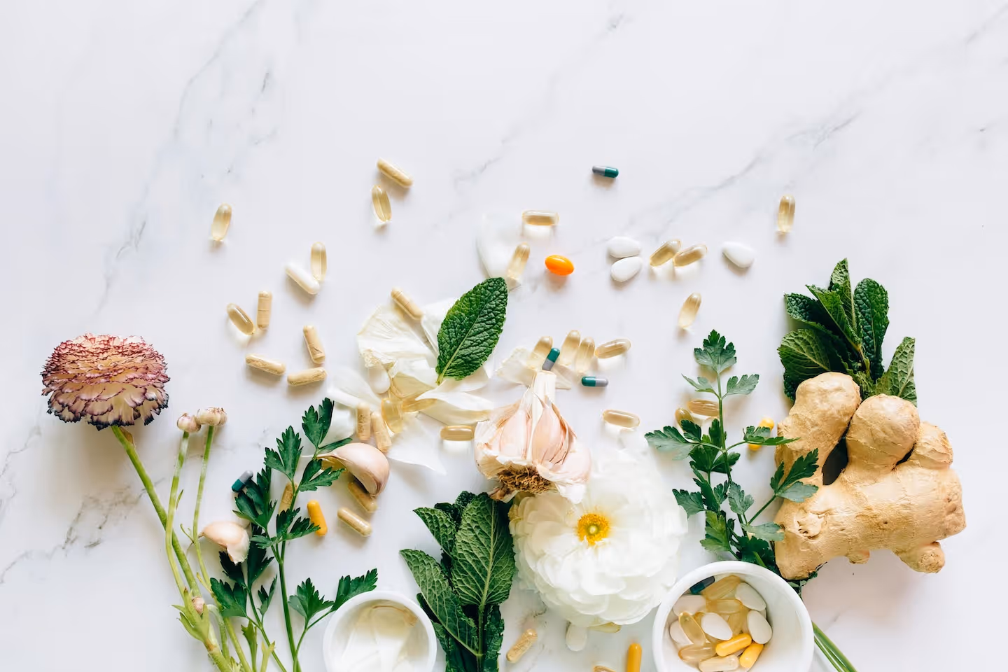 Various herbs, garlic, ginger, white and purple flowers, and capsules scattered on a white marble surface with two small bowls containing pills.