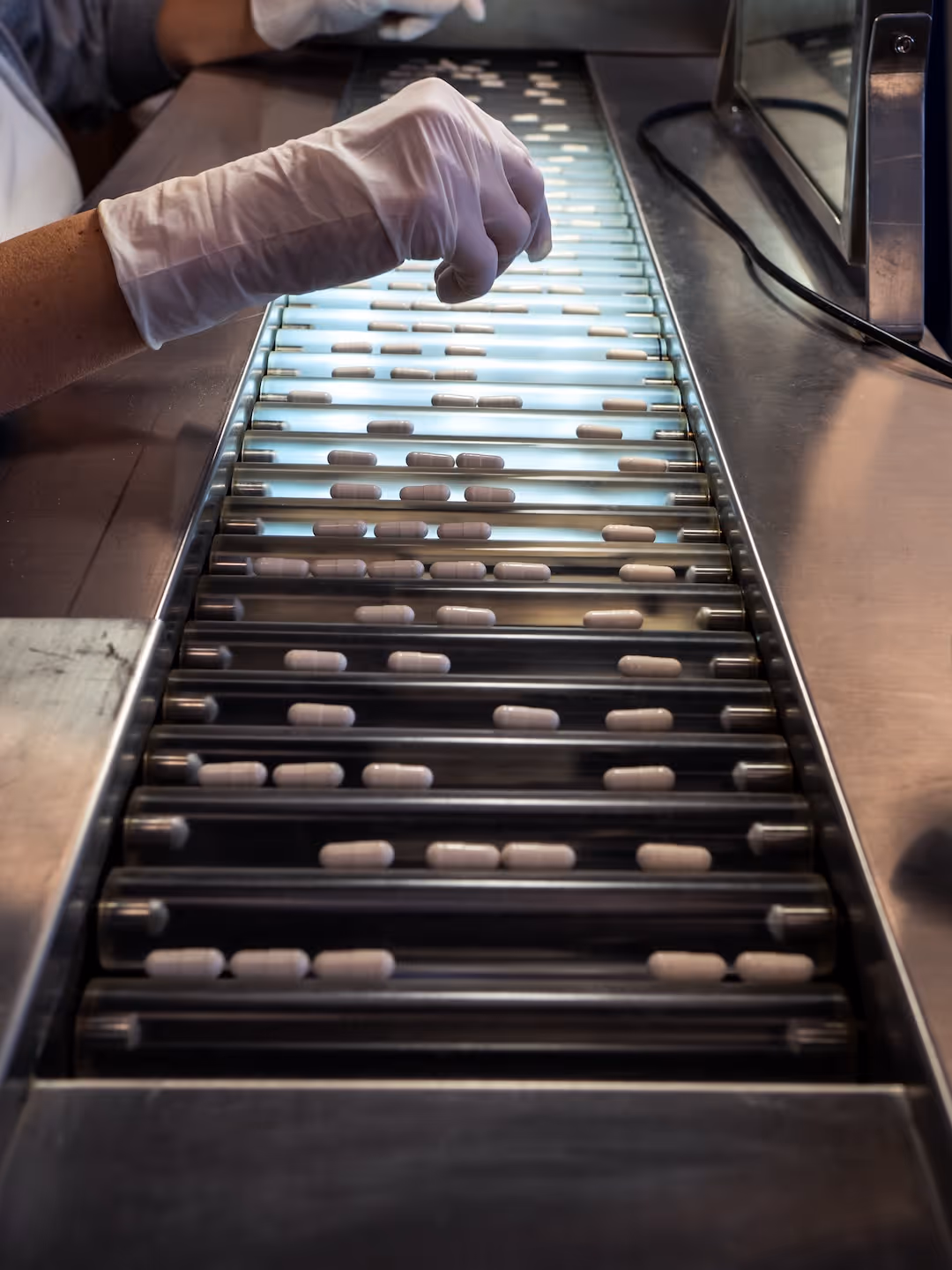 Person wearing white gloves sorting white capsules on a pharmaceutical production conveyor belt.