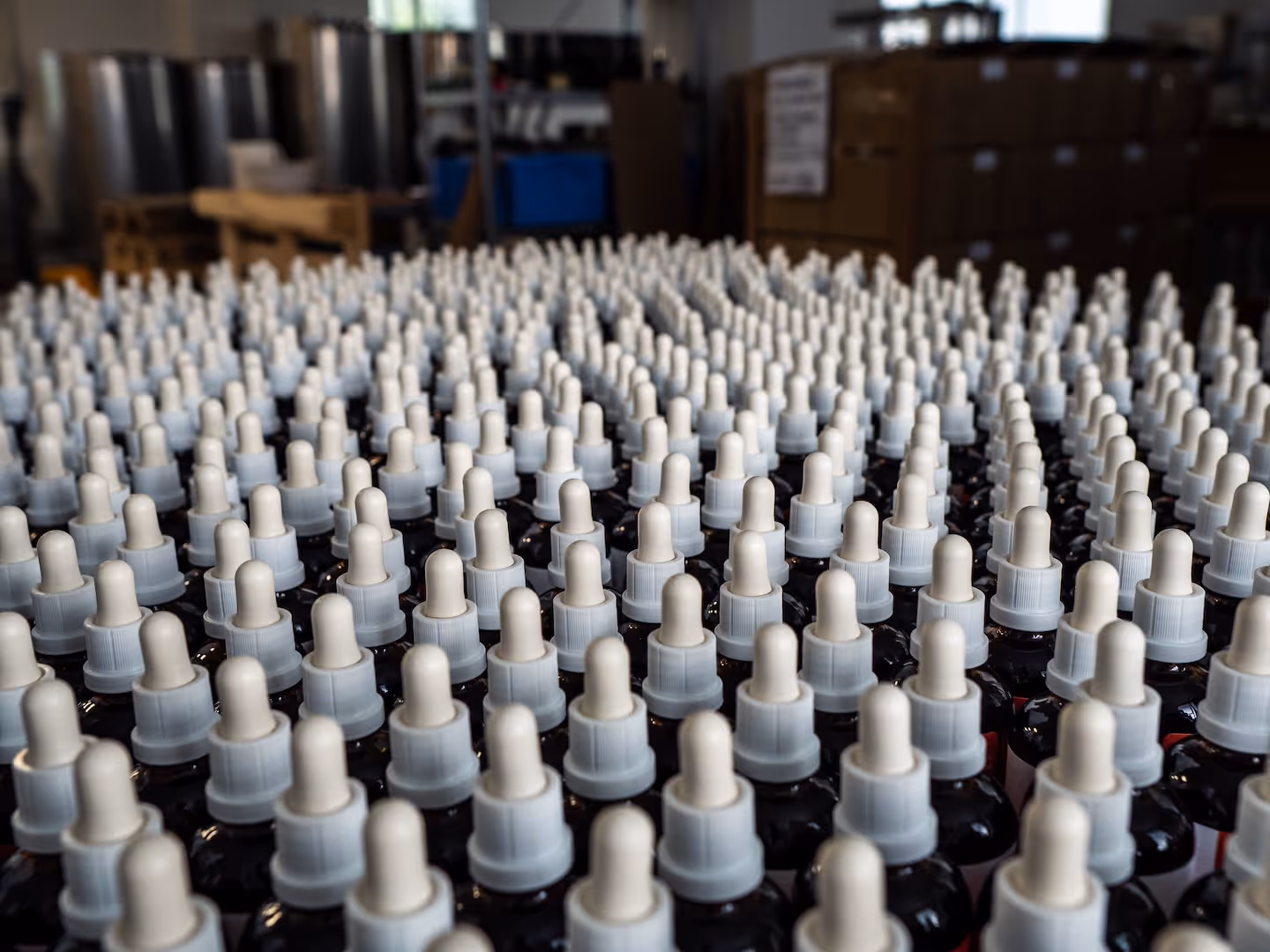 Rows of dark dropper bottles with white caps assembled on a surface in a warehouse or production facility.