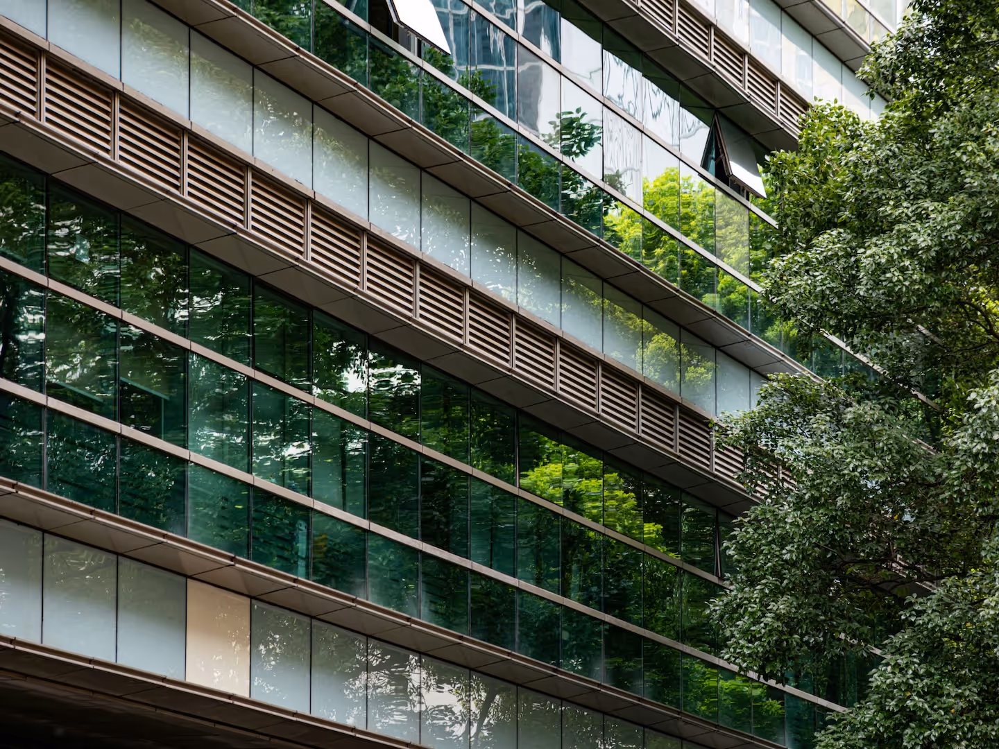 Modern building facade with green-tinted glass windows reflecting surrounding trees and greenery.