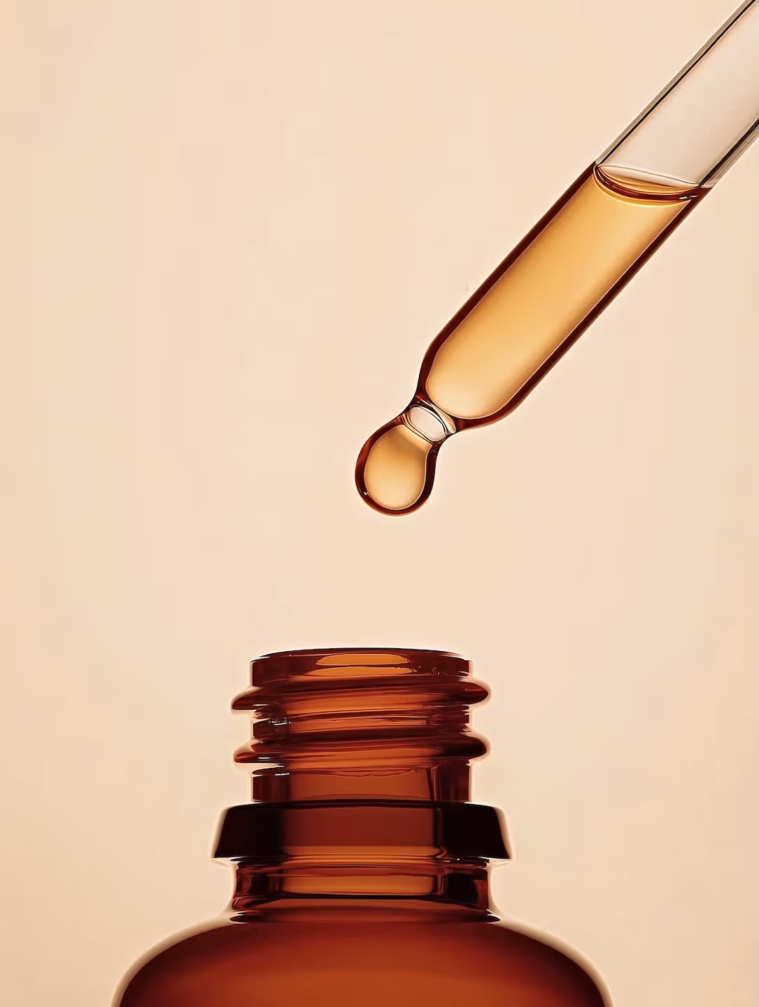 Close-up of a dropper with a golden liquid drop above the opening of a brown glass bottle.