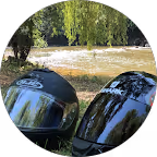 Deux casques de moto noirs posés sur une surface près d'un arbre et d'un plan d'eau, avec de la végétation verte.
