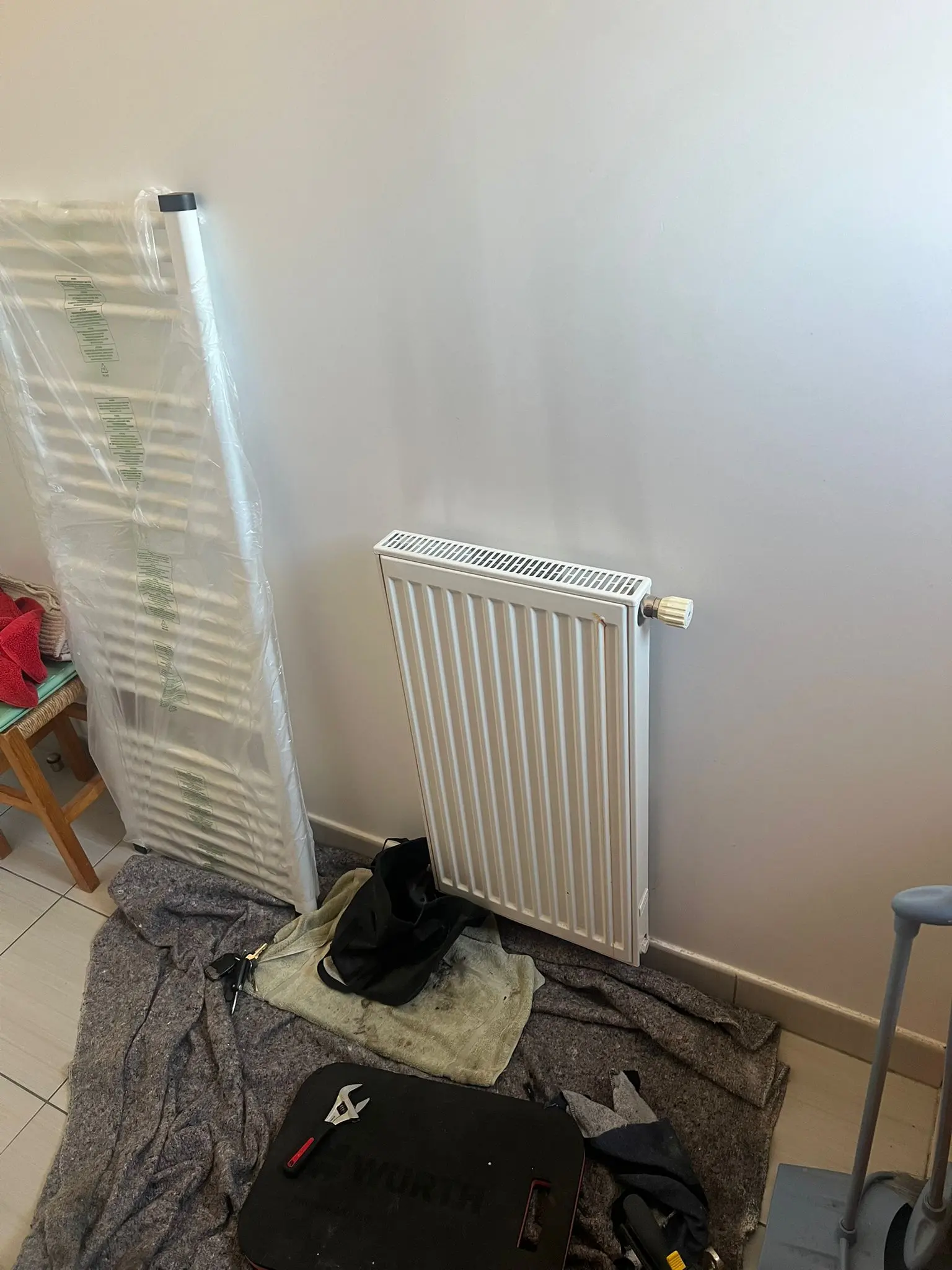 White wall-mounted radiator with a thermostat knob installed on a pale wall, surrounded by tools and protective cloth on the floor.