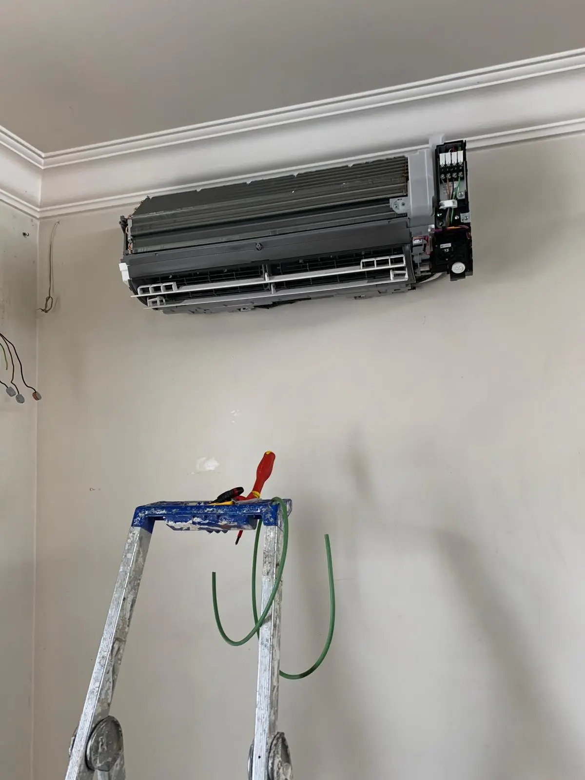Unité de climatisation murale partiellement installée avec câblage apparent au-dessus d'une échelle sur laquelle sont rangés des outils et des câbles verts