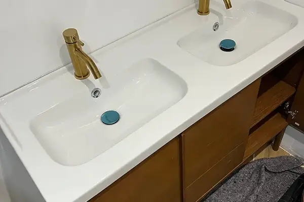 Double vasque blanche avec robinetterie dorée, fixée sur un meuble-lavabo en bois.