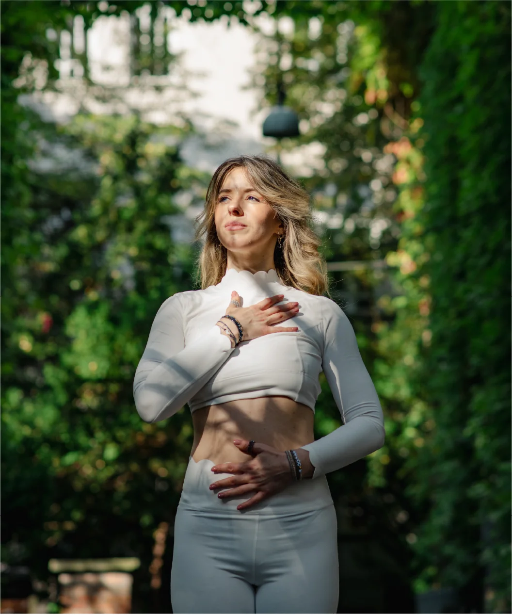 Woman in white activewear standing outdoors with one hand on her chest and the other on her abdomen, surrounded by greenery.