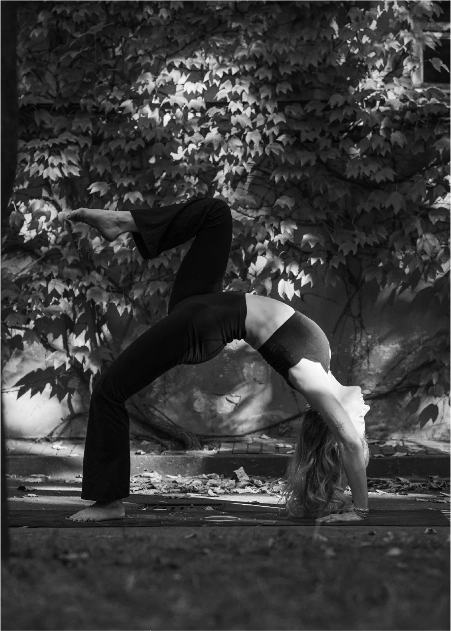 Person performing a yoga bridge pose with one leg raised outdoors in front of a leafy wall.
