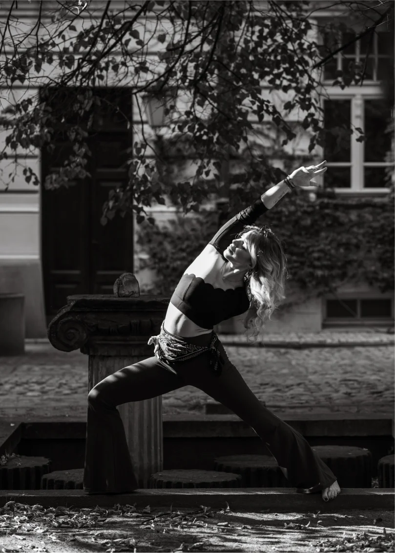 Woman practicing yoga outdoors in a warrior pose with one arm raised, under tree branches in front of a building.