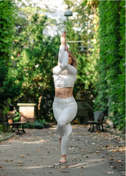 Woman in white activewear performing a yoga pose on one leg in a leafy outdoor pathway.