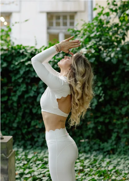 Woman in white workout clothes with long blonde hair practicing yoga stretch outdoors in front of green ivy-covered wall.