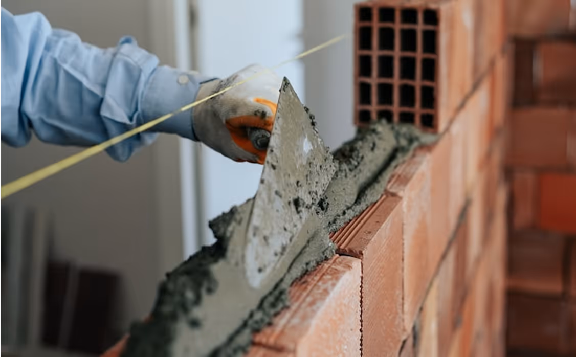 Worker spreading cement mortar on red bricks while building a wall using a trowel and string line.