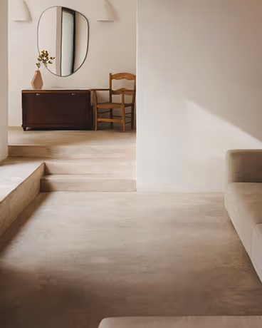 Minimalist interior with beige sofa, wooden chair, dark cabinet, oval mirror, and vase with flowers.