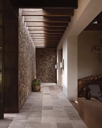 Modern stone hallway with wooden ceiling beams, potted plant, and doorway with wall lights.