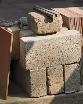 Stack of rough concrete bricks with metal tools and tiles nearby in outdoor light.