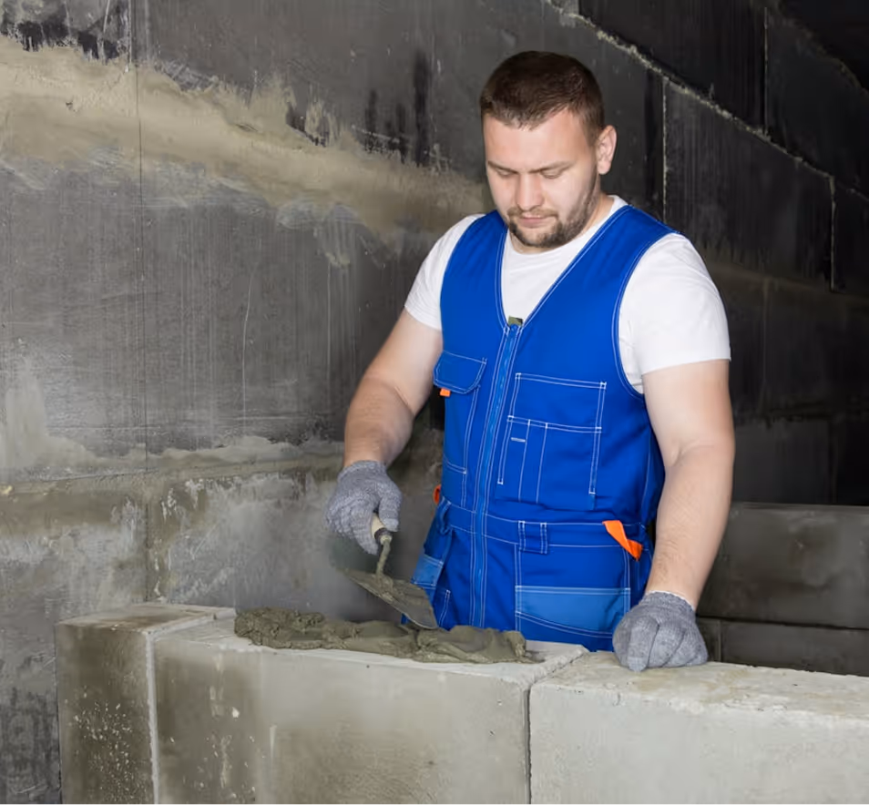 Construction worker in blue vest spreading mortar on concrete blocks with a trowel.