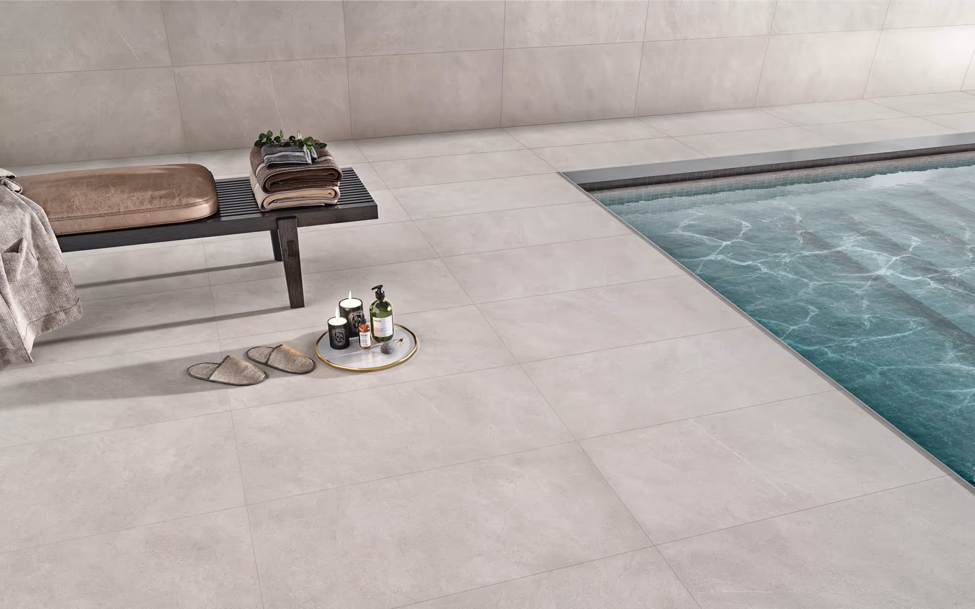 Indoor poolside area with light grey tiles, bench, folded towels, slippers, and spa items on a tray.