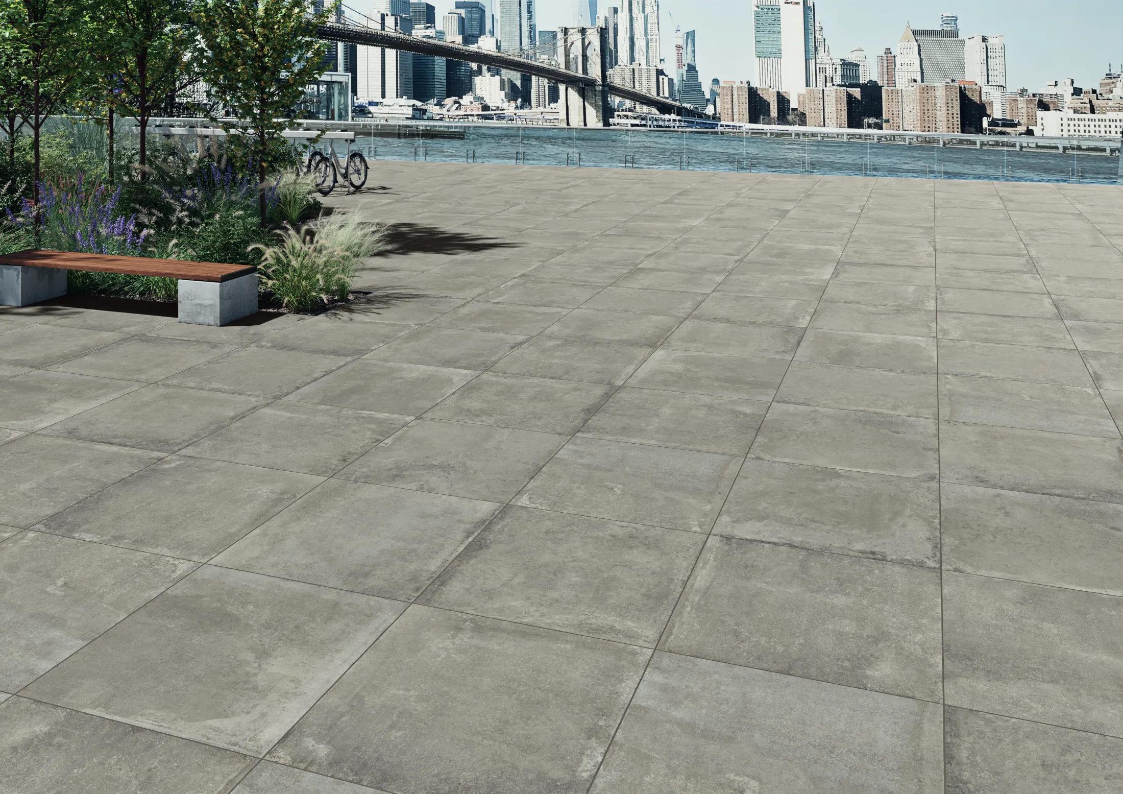 Concrete tiled plaza with bench, plants, and bike near riverfront skyline and bridge in background.