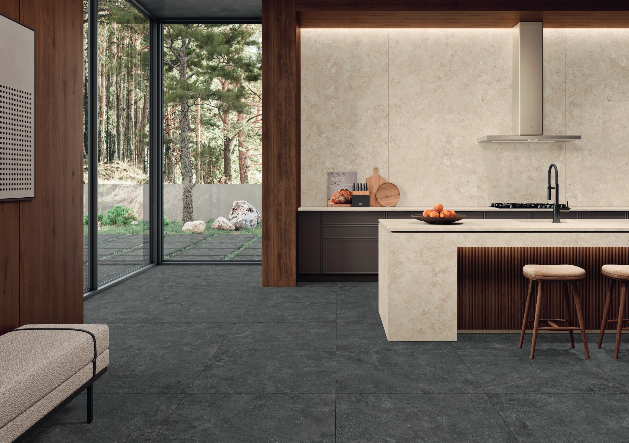 Modern kitchen with beige island, dark floor tiles, wooden accents, large windows, and outdoor forest view.