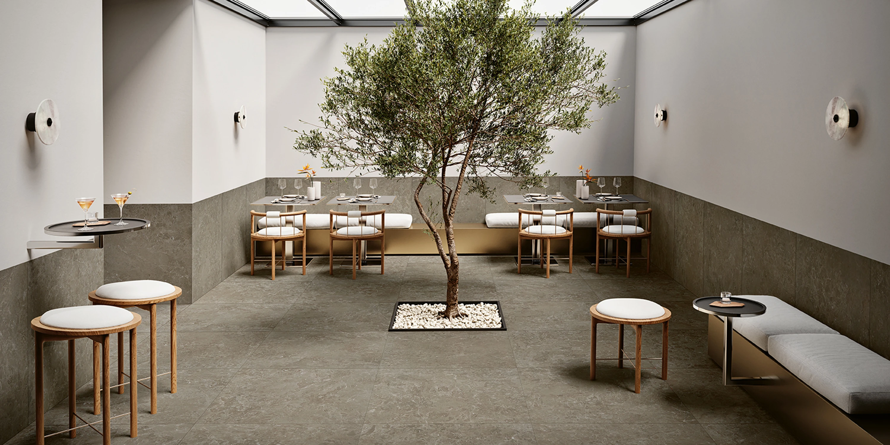 Modern dining area with central tree, gray stone flooring, wooden stools, and tables set with glassware.